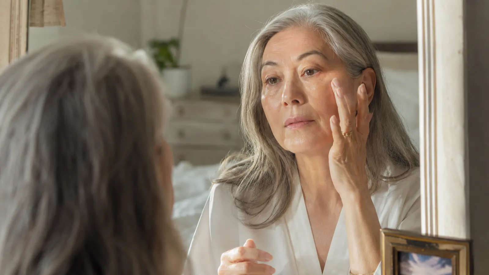 Woman in her sixties applying serum to her face in natural daylight by a bedroom mirror, calm expression, skin looking smooth and even