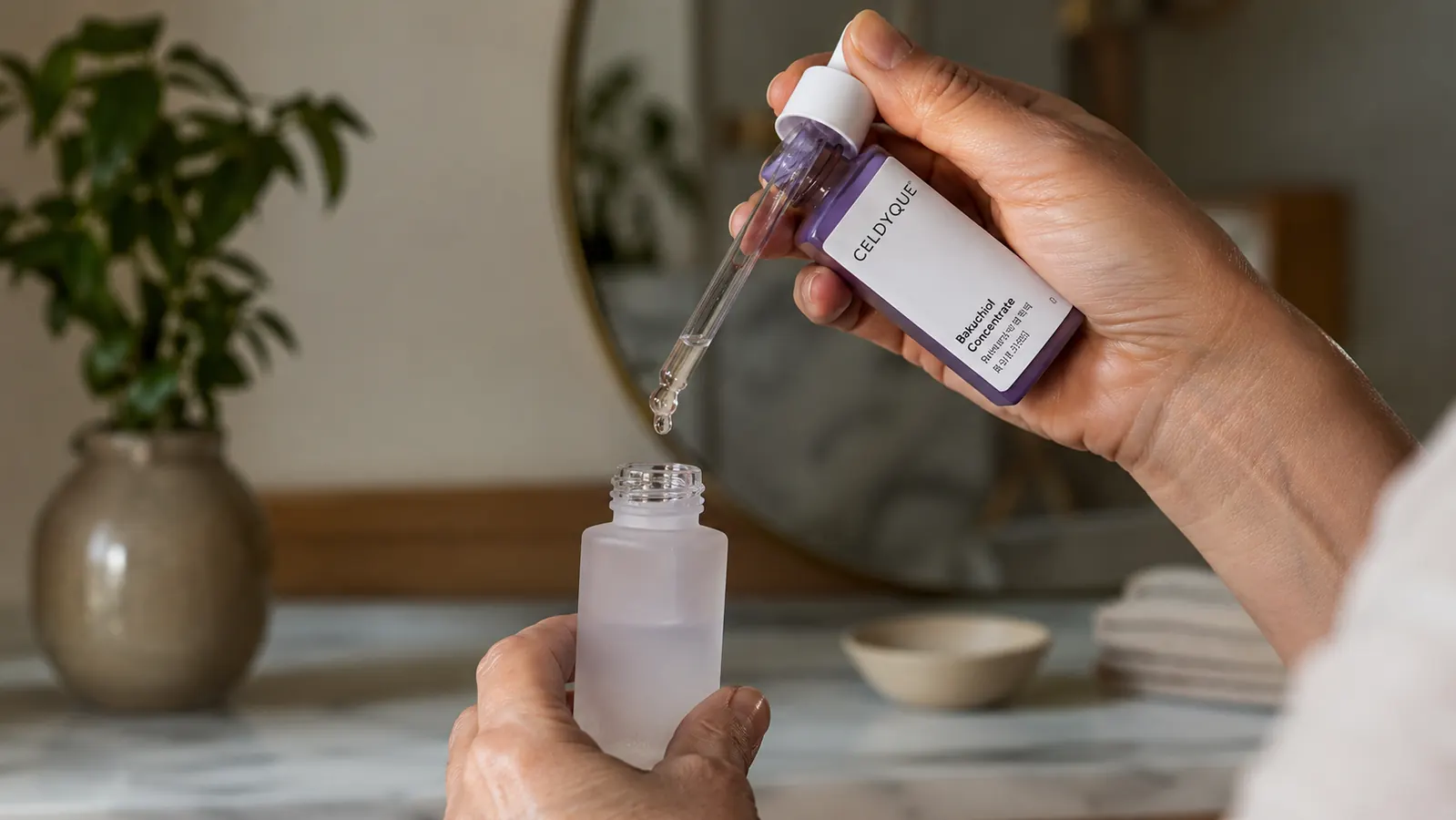 Hand holding a dropper of concentrated serum above a small mixing bottle of moisturizer on a bathroom counter