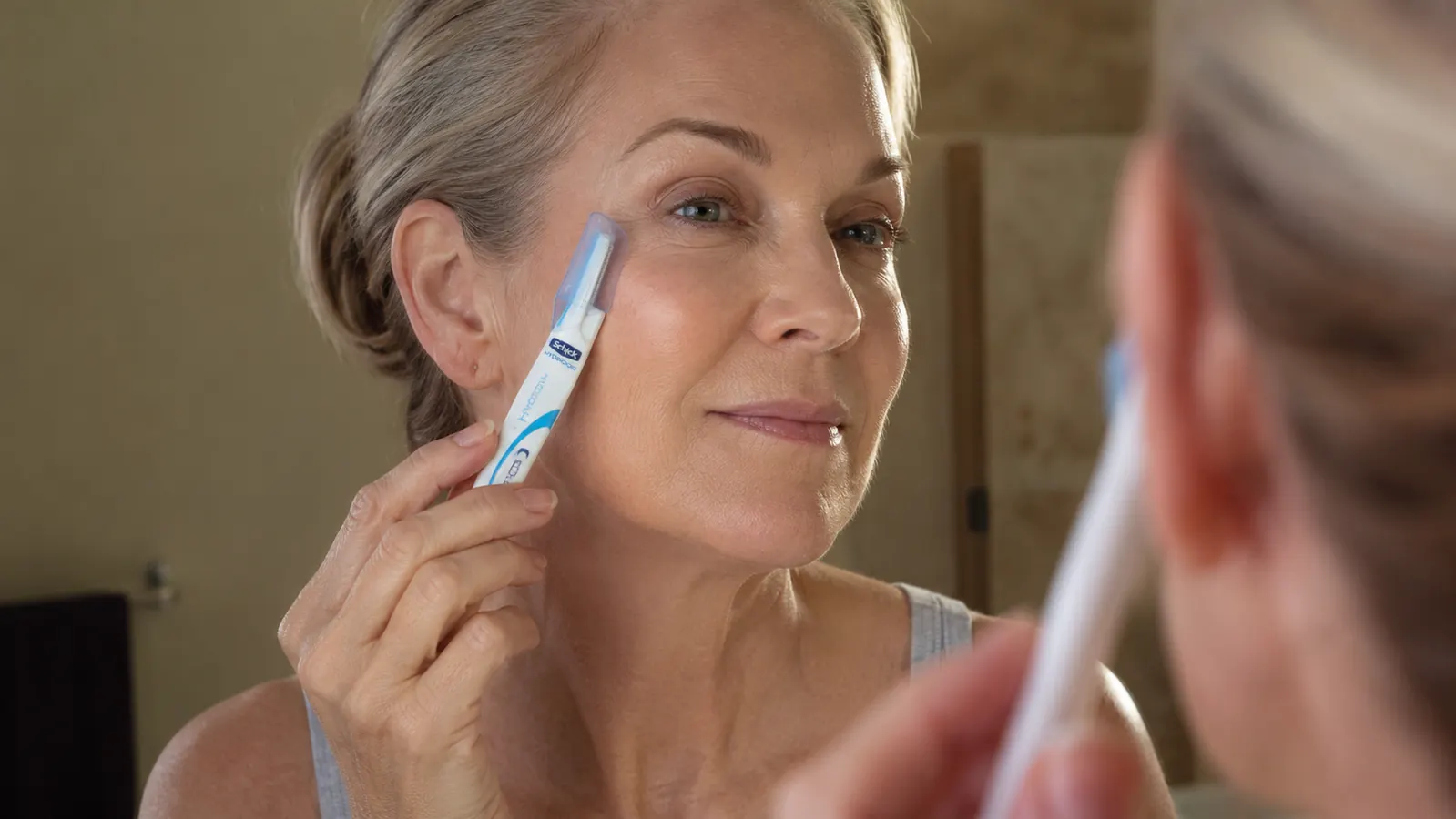 Hand holding a small dermaplaning razor tool near a cheek at a bathroom mirror