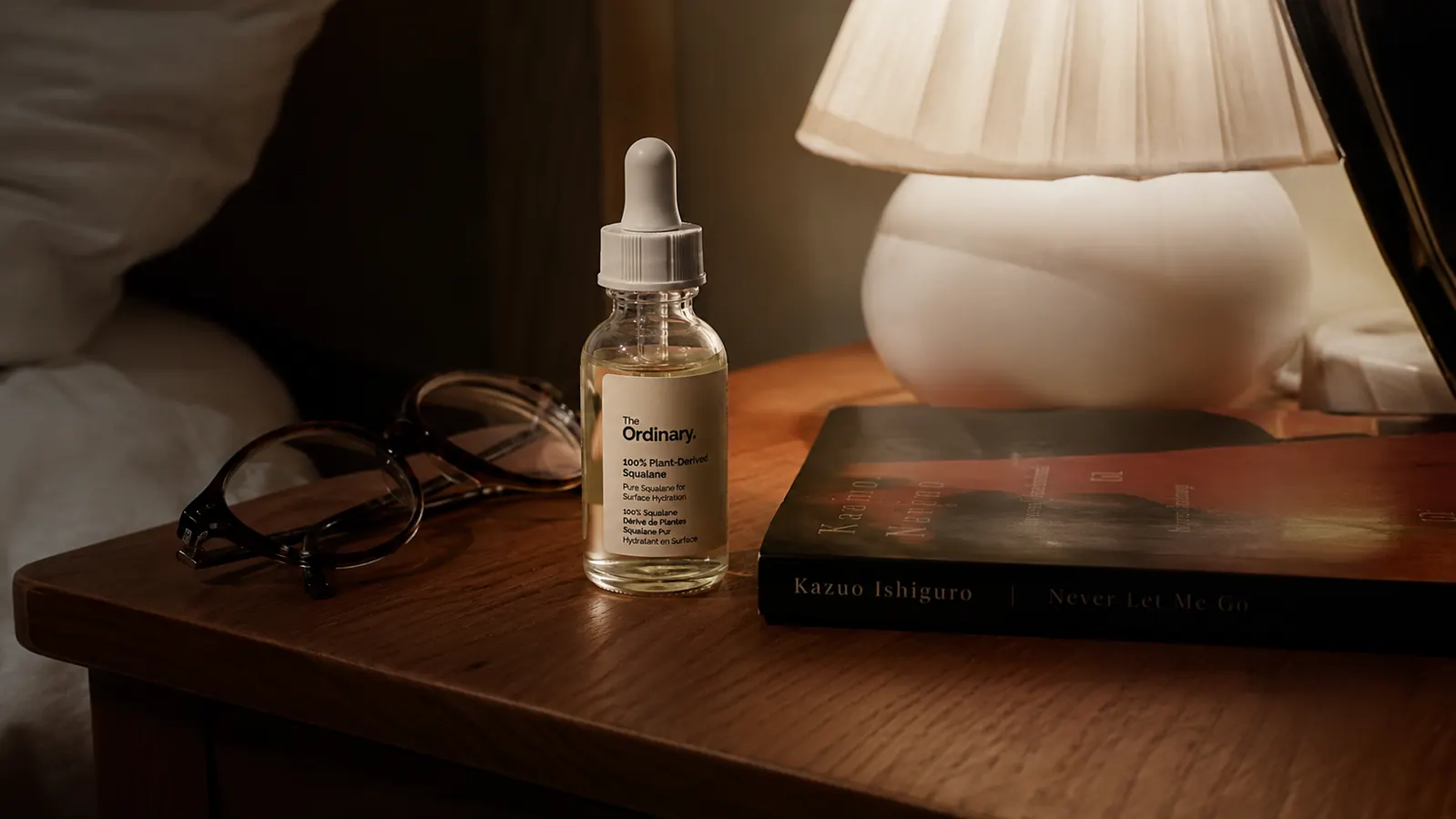 Bottle of squalane facial oil sitting on a nightstand next to a folded hand towel and reading glasses